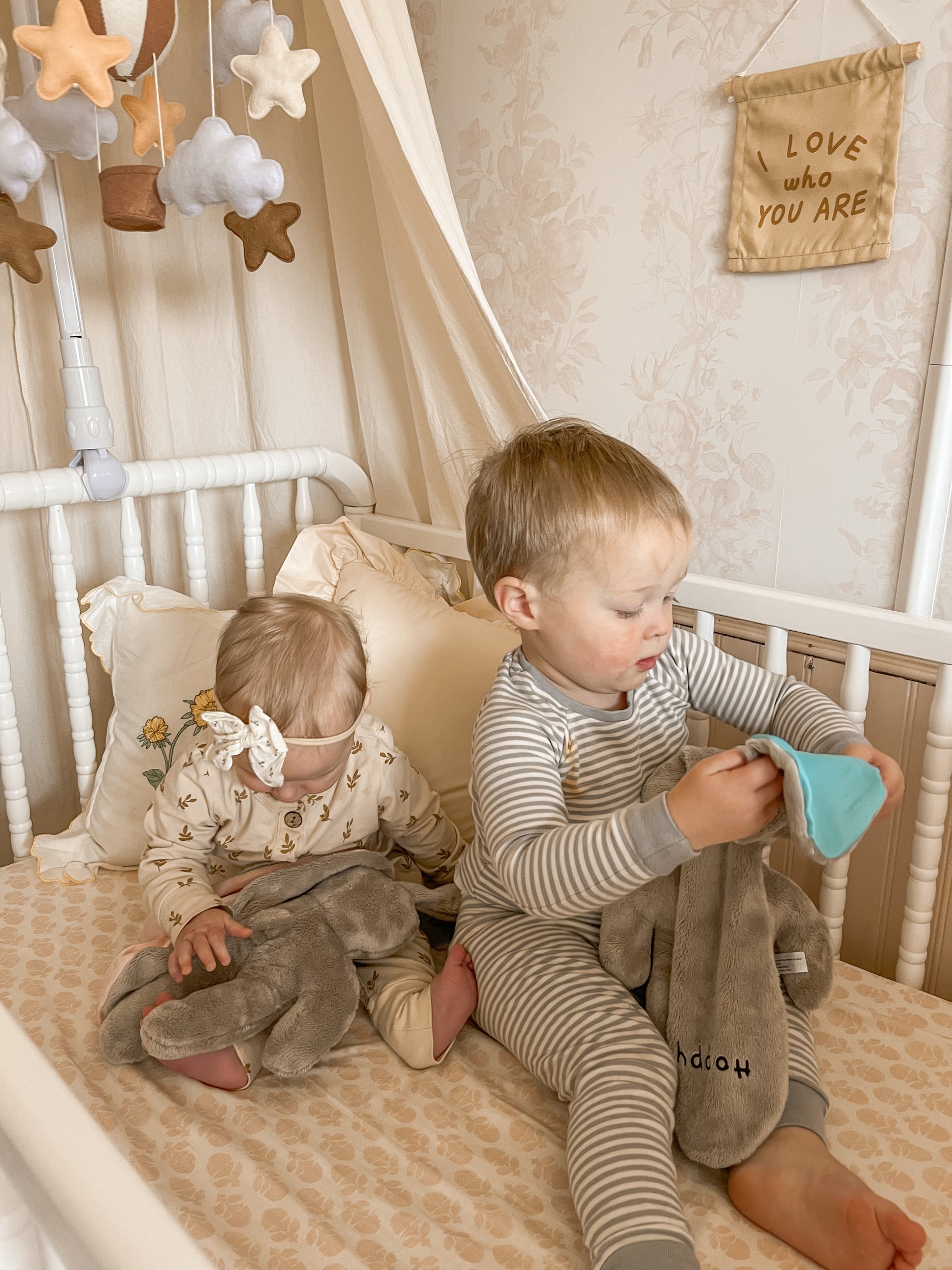 Toddler and baby holding soft Hoppy lovey bunnies in a neutral nursery, providing comfort and security during naps and quiet moments.